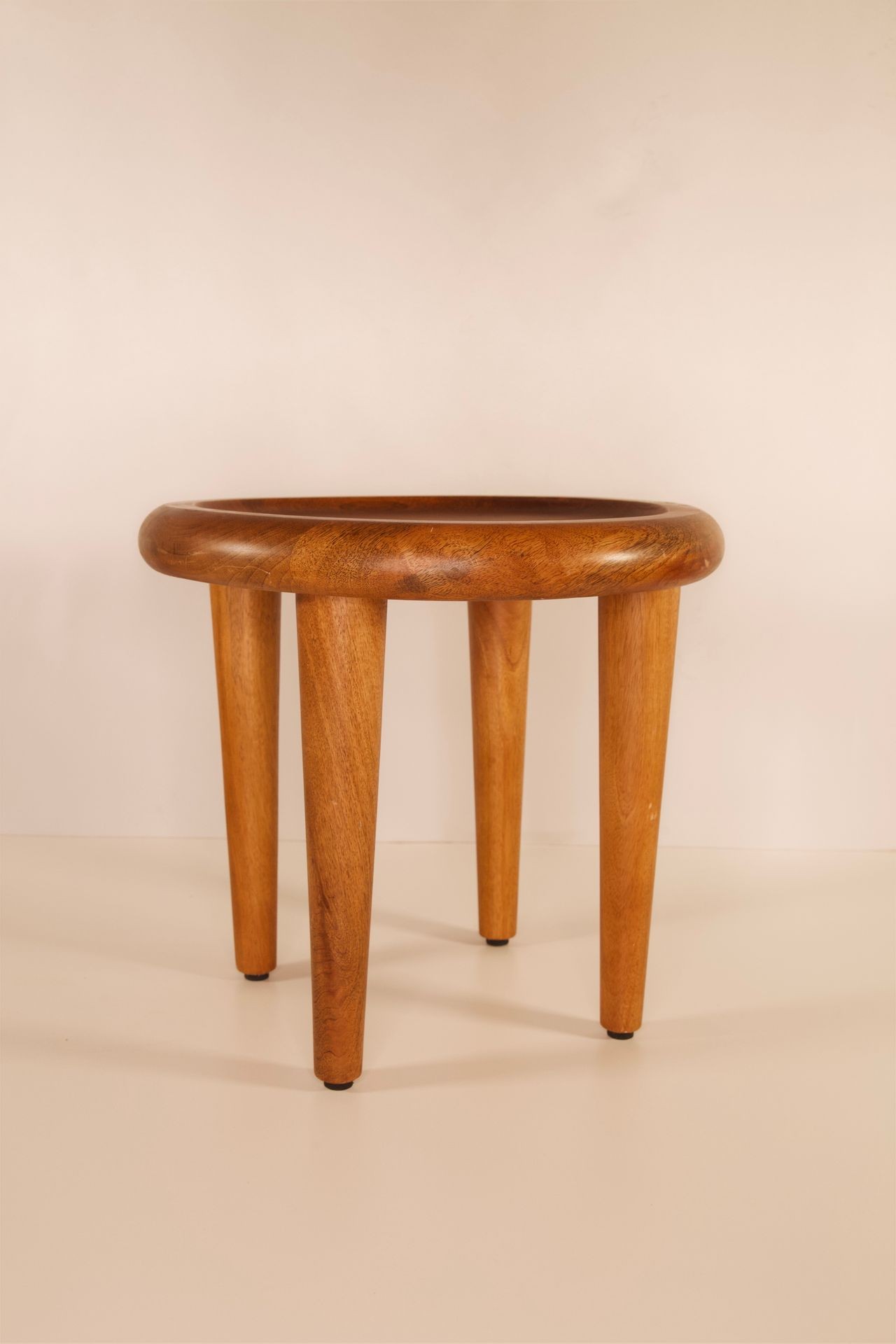 A minimalist wooden stool with four legs and a rounded seat, displayed against a plain beige background.