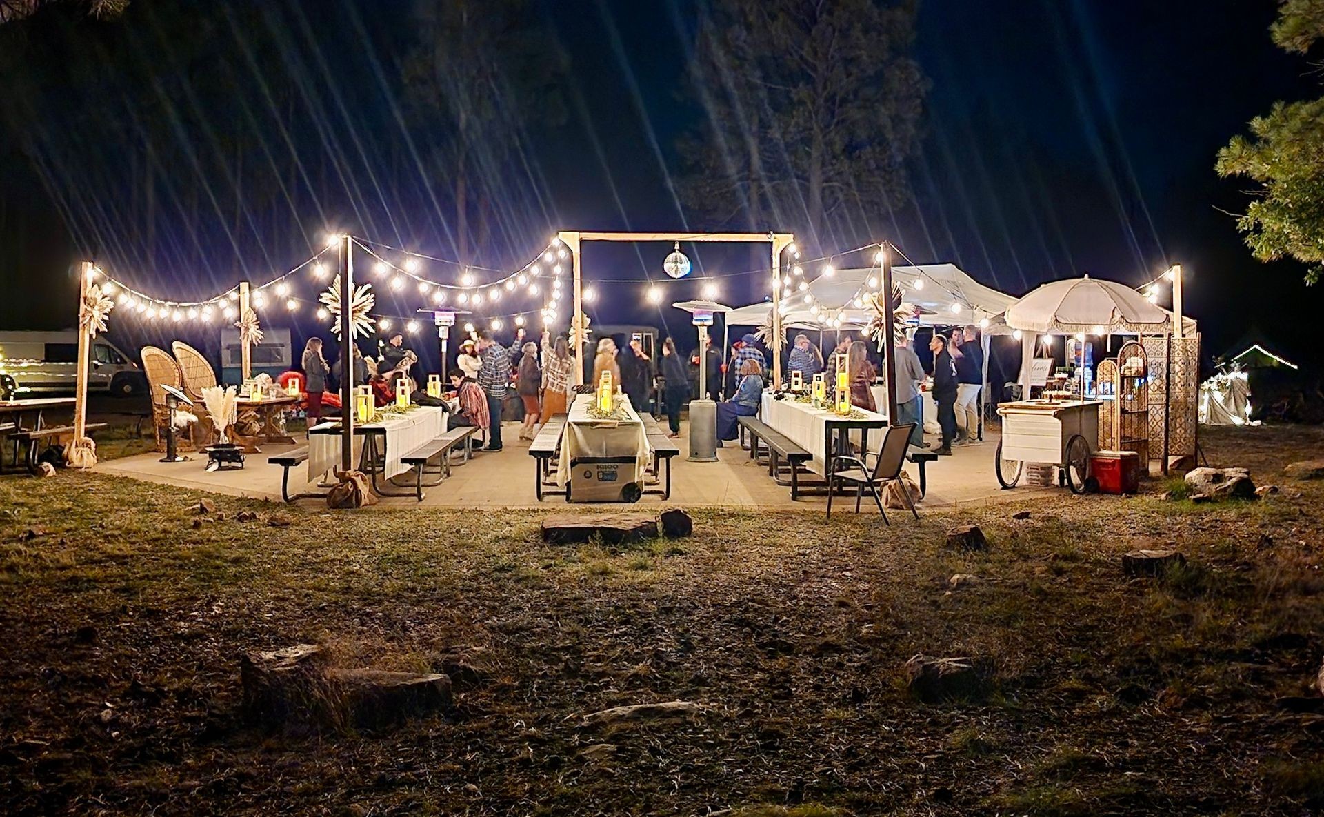 Outdoor night event with string lights, decorated tables, and people socializing under a wooden canopy.