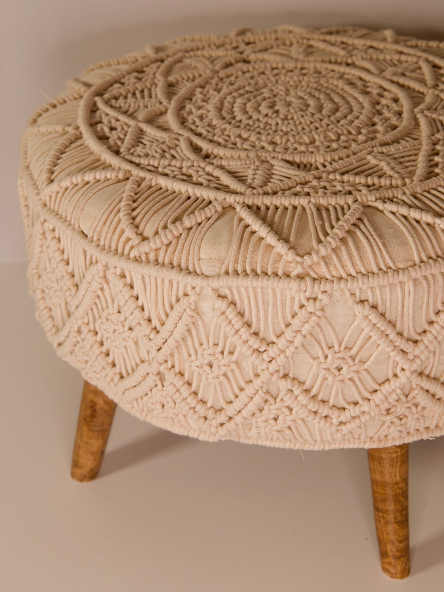 Close-up of a beige round ottoman with intricate macramé design and wooden legs.