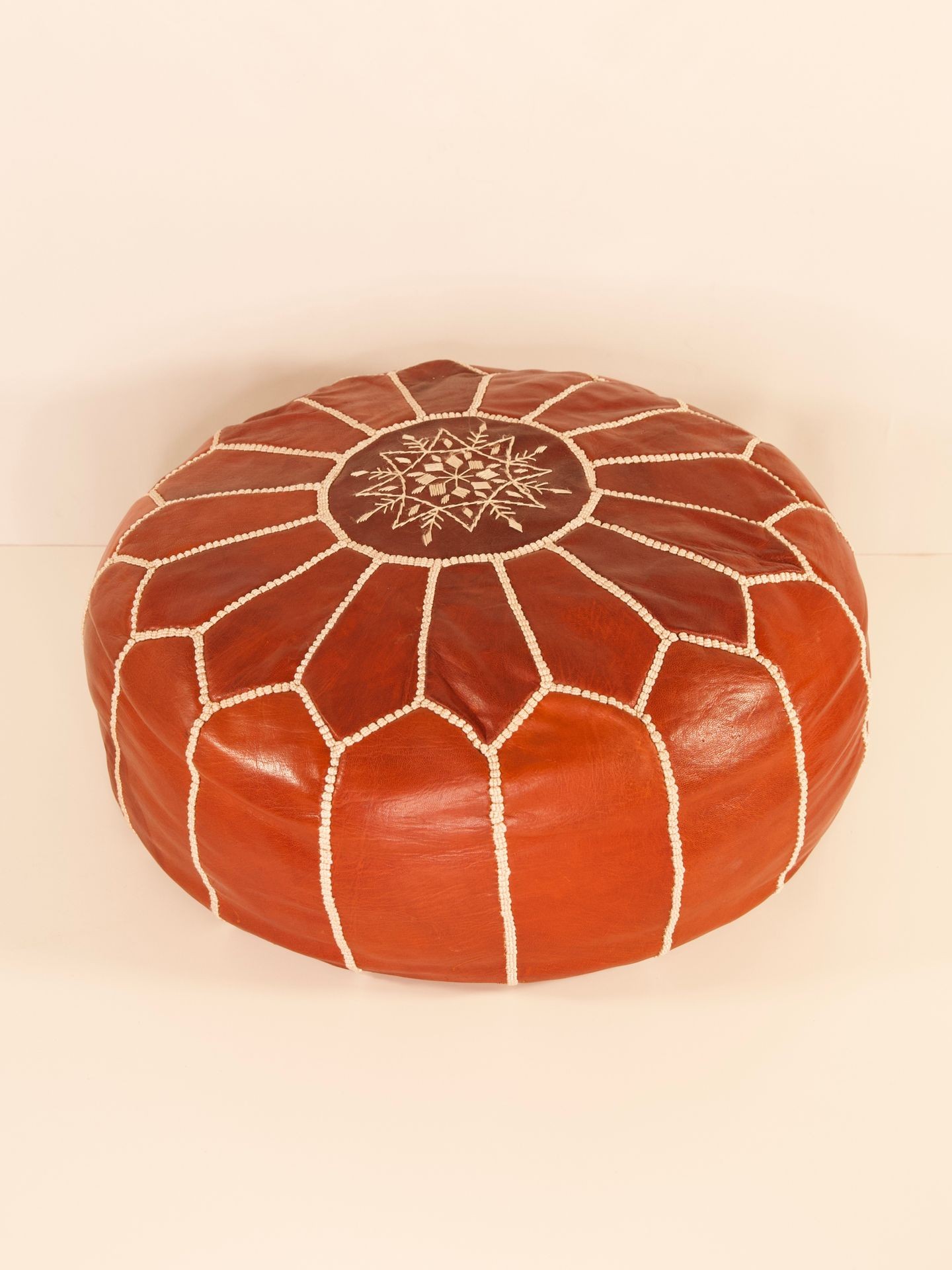 Round, brown leather pouf with white stitching and intricate patterned design on top.