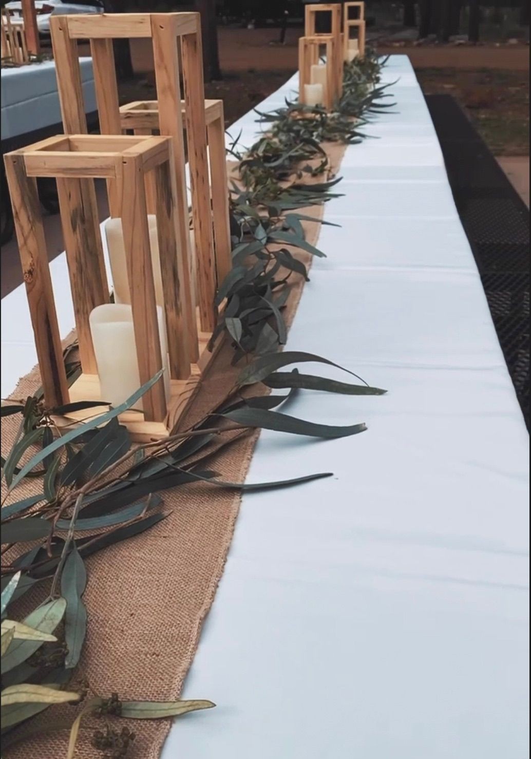 Outdoor table setting with rustic wooden candle holders and greenery on a burlap table runner.