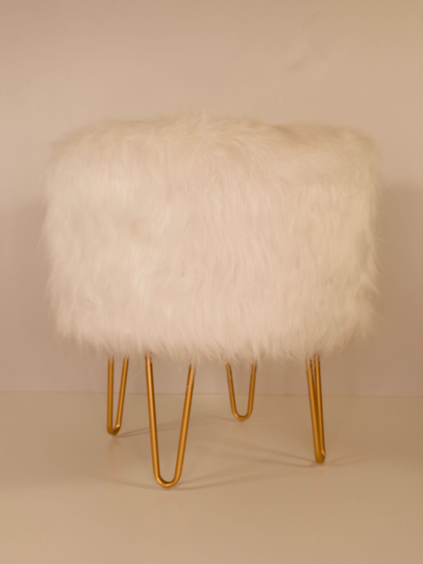 White faux fur ottoman with gold hairpin legs, placed in a minimalistic indoor setting.